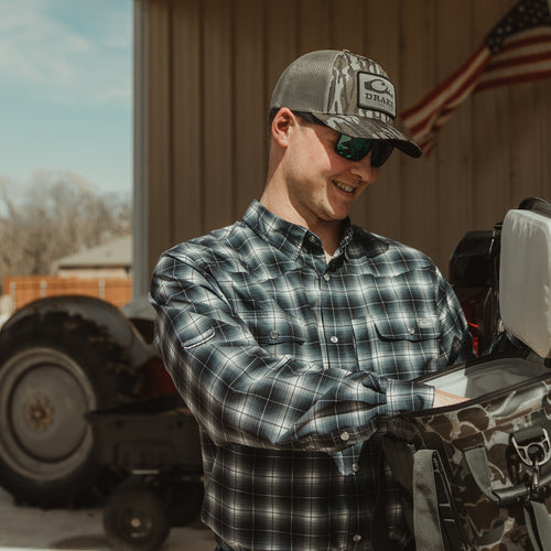 A man wearing a Cinco Ranch Western Fall Plaid Button-Down Shirt, with hat and sunglasses, examining a bag outdoors.
