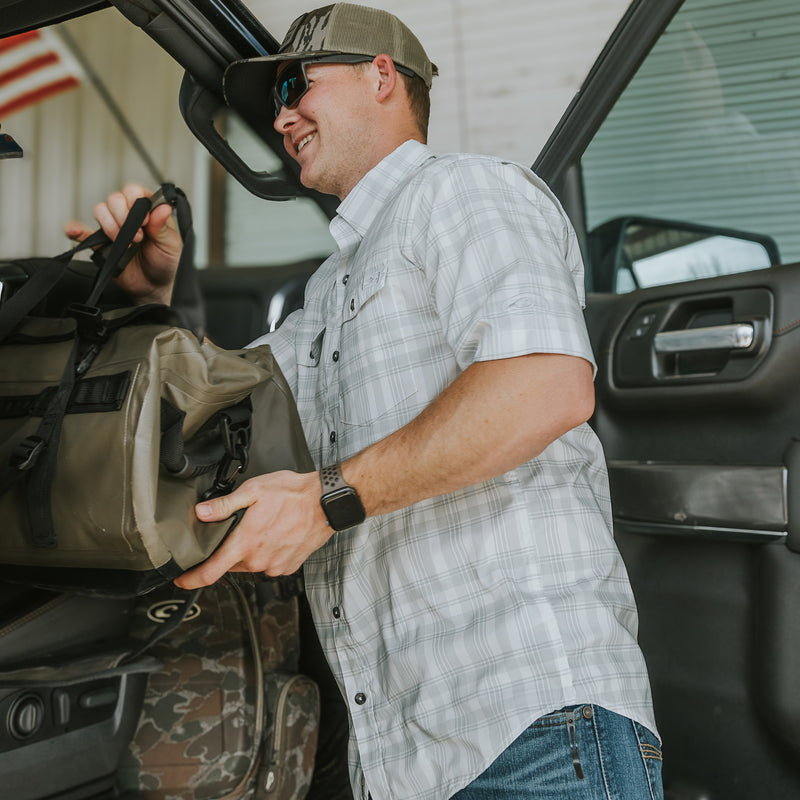 A man in sunglasses and a hat holding a bag near a vehicle, showcasing the Frat Faded Plaid Button-Down Short Sleeve Shirt from Purewhitelaneshop. Features UPF 30, moisture-wicking, and hidden collar.