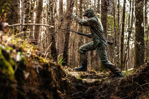 Man in camouflage showcasing Throwback 80 Field Pant with reinforced knees, antique brass riveted pockets, and YKK zippered fly in a woodland setting.