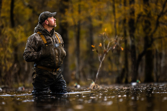 A bearded man wearing the LST BMF Jacket with Primaloft scans the sky in a flooded forest, highlighting its water-resistant, lightweight design for active hunting.