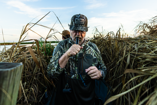 Hunter in camouflage using a duck call, showcasing the EST Performance Half-Zip Hoodie with ergonomic thumbholes and water-resistant pocket in a wetland hunting setup.