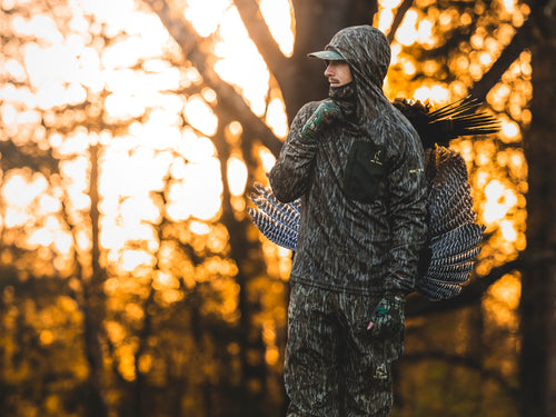 Man in camouflage Early Bird Gator Hoodie With Insect Repellent, featuring feathers, forest backdrop, showcasing hunting gear's protective and versatile design.