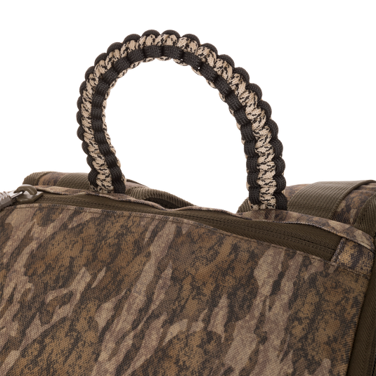 Close-up of the Tree Hugger Timber Pack showing its brown-and-green camo pattern, zippered pocket, and braided paracord carry-handle loop, emphasizing durability and organization.