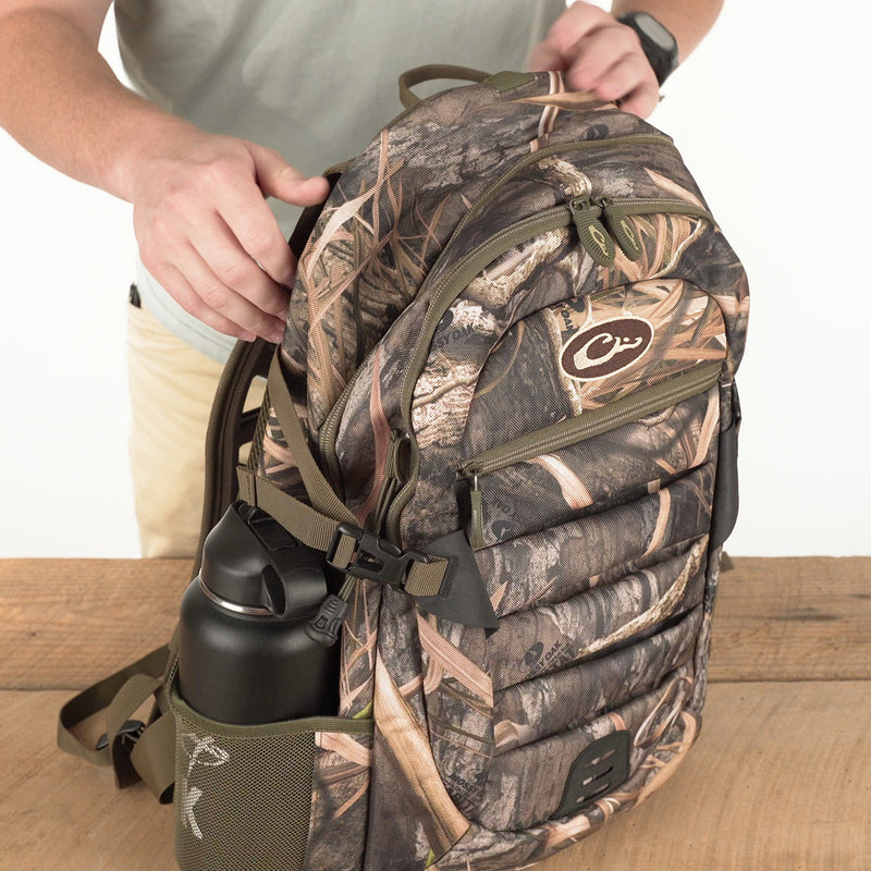 Youth Camo Daypack on a wooden table, featuring a camo pattern, side mesh pocket with a water bottle, and adjustable straps, ideal for casual or hunting use.