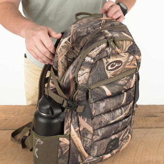 Youth Camo Daypack on a wooden table, featuring a camo pattern, side mesh pocket with a water bottle, and adjustable straps, ideal for casual or hunting use.