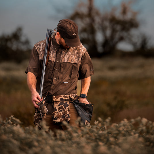 Hunter in camo gear with a shotgun and quail, showcasing the Drake Wingshooter's Dove Belt's spacious game bag and functional design for organized hunting convenience.