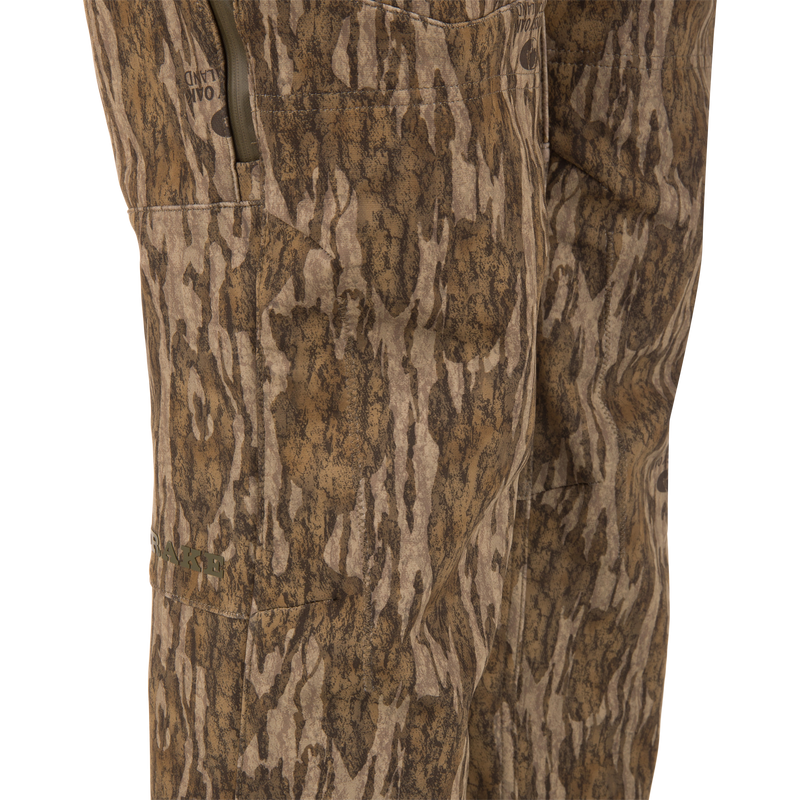 Close-up of MST CommandXT Softshell Pant in wood-grain camo, featuring a zippered side pocket, OAKLAND branding, and DRAKE logo, highlighting mobility and protection.