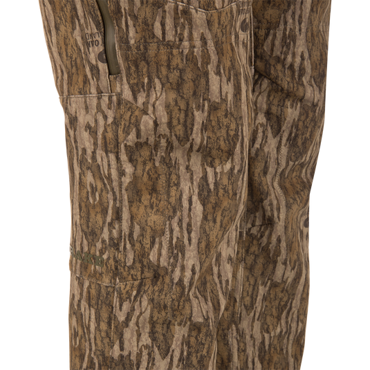 Close-up of MST CommandXT Softshell Pant in wood-grain camo, featuring a zippered side pocket, OAKLAND branding, and DRAKE logo, highlighting mobility and protection.