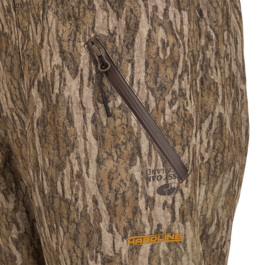 LST Hardline Insulated Bib with camo print, featuring a waterproof zipper pocket, Mossy Oak Bottomland logo, and HARDLINE in orange, highlighting durability and weather protection.
