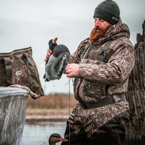 A man in a camouflage hoodie holding ducks in water, showcasing the MST Endurance Soft Shell Hoodie by Purewhitelaneshop. Features Magnattach™ pocket, zipped lower pockets, mesh-lined sleeves, and fleece-lined hood.