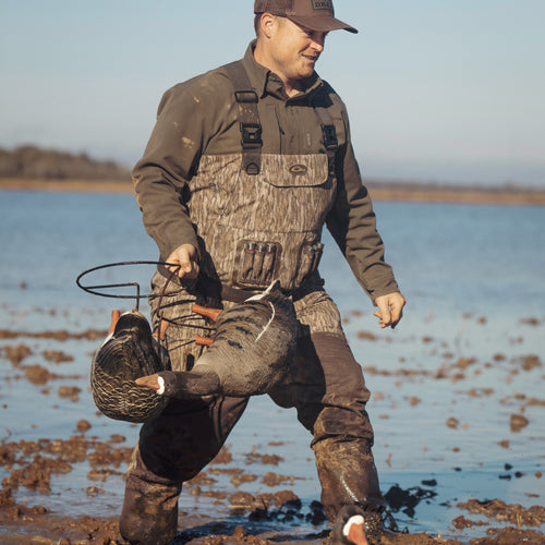 A man in hunting gear carrying decoys in mud, showcasing the Insulated Guardian Elite Vanguard Breathable Waders by Purewhitelaneshop. Waterproof, windproof, with LokDown insulation and Thinsulate boots for warmth and comfort.