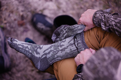 A person outdoors pulls up a Midweight Merino Camo Mid Calf Hunting Sock over their foot, showcasing its camo pattern and durable design.
