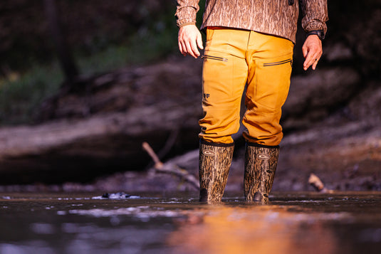 Person waist-deep in water wearing MST CommandXT Softshell Pant with waterproof features and camouflage wading boots, showcasing mobility and protection for demanding outdoor activities.