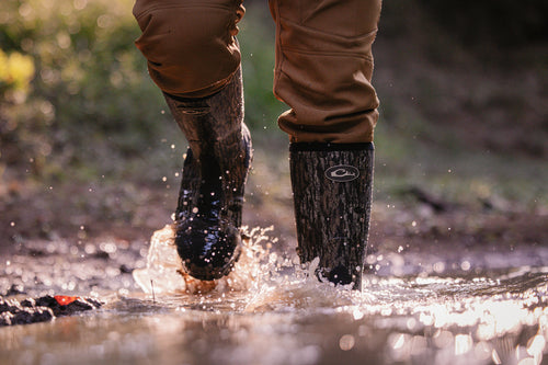 Person walking through puddle in camouflage EST Uninsulated 16 Hybrid Mudder Boot 3.0, showcasing water-resistant durability and comfort for rugged environments.