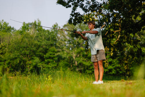 Young man casting a fishing rod, wearing the Drake Wingshooter Trey Cross Hatch Short Sleeve Shirt, showcasing its lightweight, moisture-wicking, and sun-protective features.