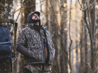 A bearded man leans against a tree in woodland, wearing the MST Offset Half Zip Hoodie, a camo-patterned, ergonomic hunting outerwear designed for comfort and functionality.