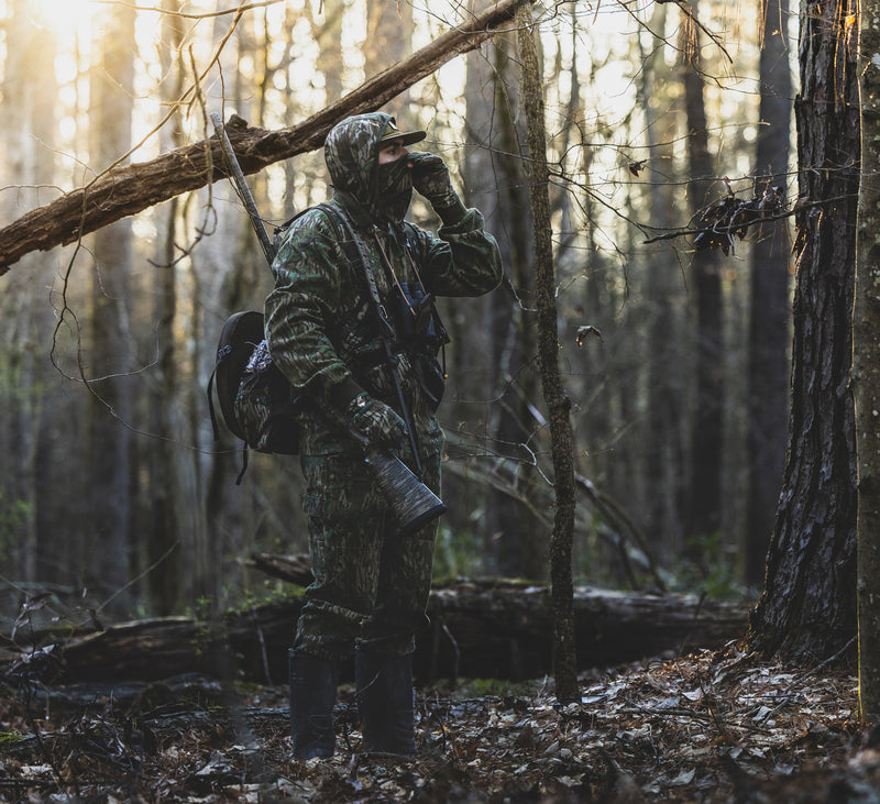 Man in camouflage wears the Throwback 80 Field Shirt Jacket, displaying its durability with reinforced elbows and riveted pockets, ideal for hunting in the woods.
