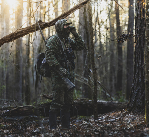 Man in camouflage wears the Throwback 80 Field Shirt Jacket, displaying its durability with reinforced elbows and riveted pockets, ideal for hunting in the woods.