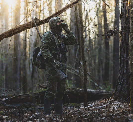 Man in camouflage wears the Throwback 80 Field Shirt Jacket, displaying its durability with reinforced elbows and riveted pockets, ideal for hunting in the woods.