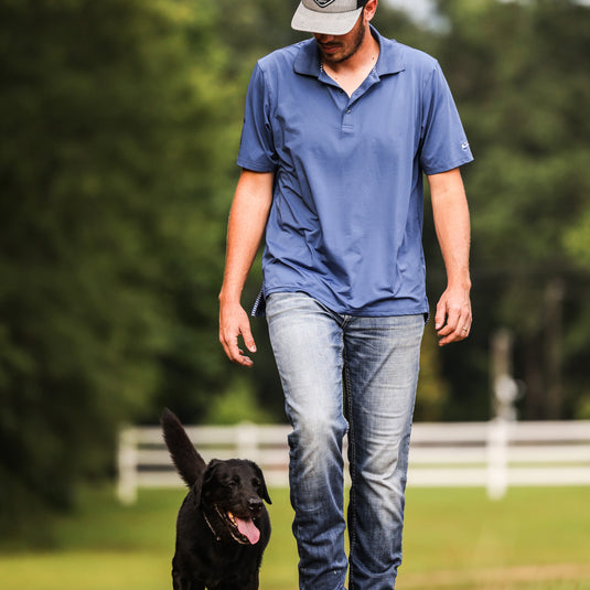 A man in a blue shirt walking a black dog outdoors. Performance Stretch Polo S/S by Purewhitelaneshop. Features 4 Way Stretch, Moisture Wicking, UPF Sun Protection, and Rib Knit Collar.