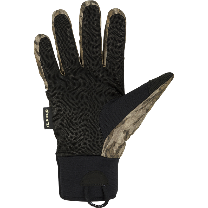 A black glove with a camouflage pattern, perfect for early season waterfowl hunting. Made with waterproof Refuge HS™ shell fabric and GORE-TEX® membrane for 100% waterproof/breathable protection. Features include a digitized goat skin leather palm and gusseted neoprene cuffs. Stay-Put™ liner system ensures no twisting or bunching. Adjustable gusset Velcro cuff closure and pull loop assist for easy use. From Purewhitelaneshop, your go-to store for high-quality hunting gear and clothing.