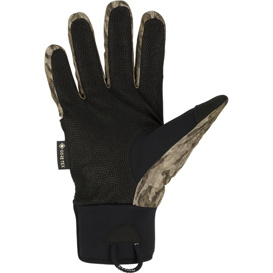 A black glove with a camouflage pattern, perfect for early season waterfowl hunting. Made with waterproof Refuge HS™ shell fabric and GORE-TEX® membrane for 100% waterproof/breathable protection. Features include a digitized goat skin leather palm and gusseted neoprene cuffs. Stay-Put™ liner system ensures no twisting or bunching. Adjustable gusset Velcro cuff closure and pull loop assist for easy use. From Purewhitelaneshop, your go-to store for high-quality hunting gear and clothing.