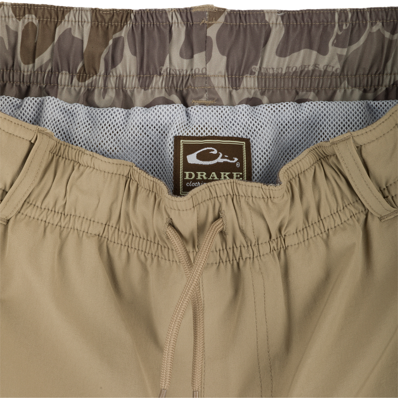 A pair of khaki Dock Swim Trunk Shorts with a label, zipper, and fabric close-ups. Made of durable nylon/spandex blend with water-resistant finish. Features elastic waist, mesh pockets, and 6