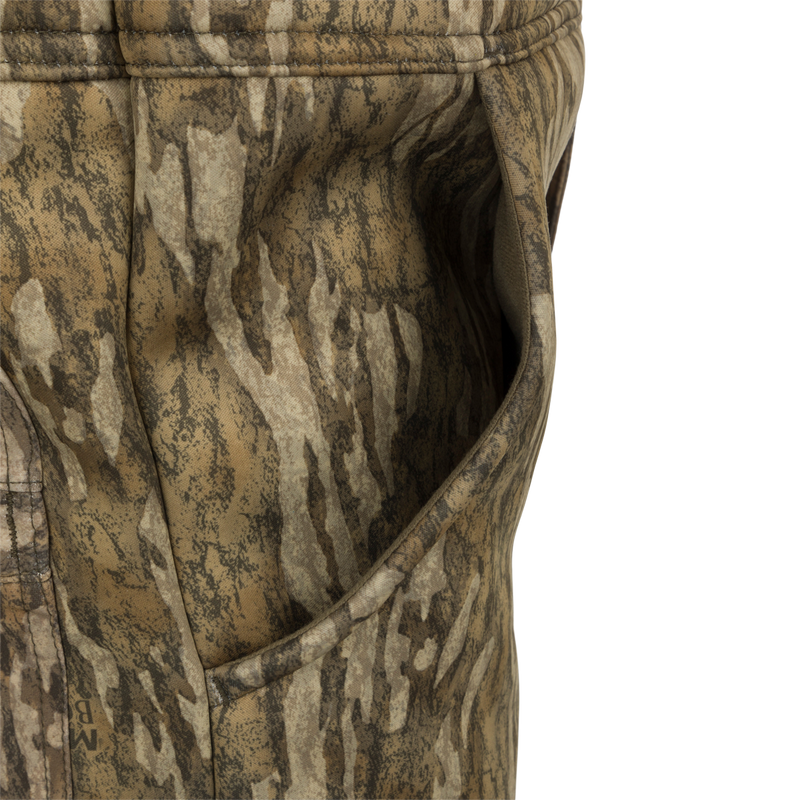 A close-up of the MST Ultimate Wader Bib, featuring a camouflage pants and jacket made of 100% polyester fleece shell with Sherpa fleece lining. The fabric has a four-way stretch, gusseted crotch, and articulated knees for easy movement. The bib also includes Sherpa-lined slash handwarmer pockets, a Magnattach™ left chest pocket, and zippered rear security pockets for storage. It seamlessly converts from a casual, over-the-boot fit to a tight, ankle-fit for wear under waders.