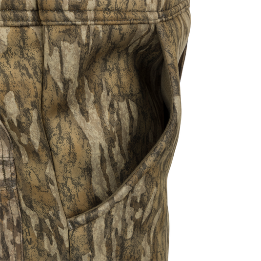 A close-up of the MST Ultimate Wader Bib, featuring a camouflage pants and jacket made of 100% polyester fleece shell with Sherpa fleece lining. The fabric has a four-way stretch, gusseted crotch, and articulated knees for easy movement. The bib also includes Sherpa-lined slash handwarmer pockets, a Magnattach™ left chest pocket, and zippered rear security pockets for storage. It seamlessly converts from a casual, over-the-boot fit to a tight, ankle-fit for wear under waders.